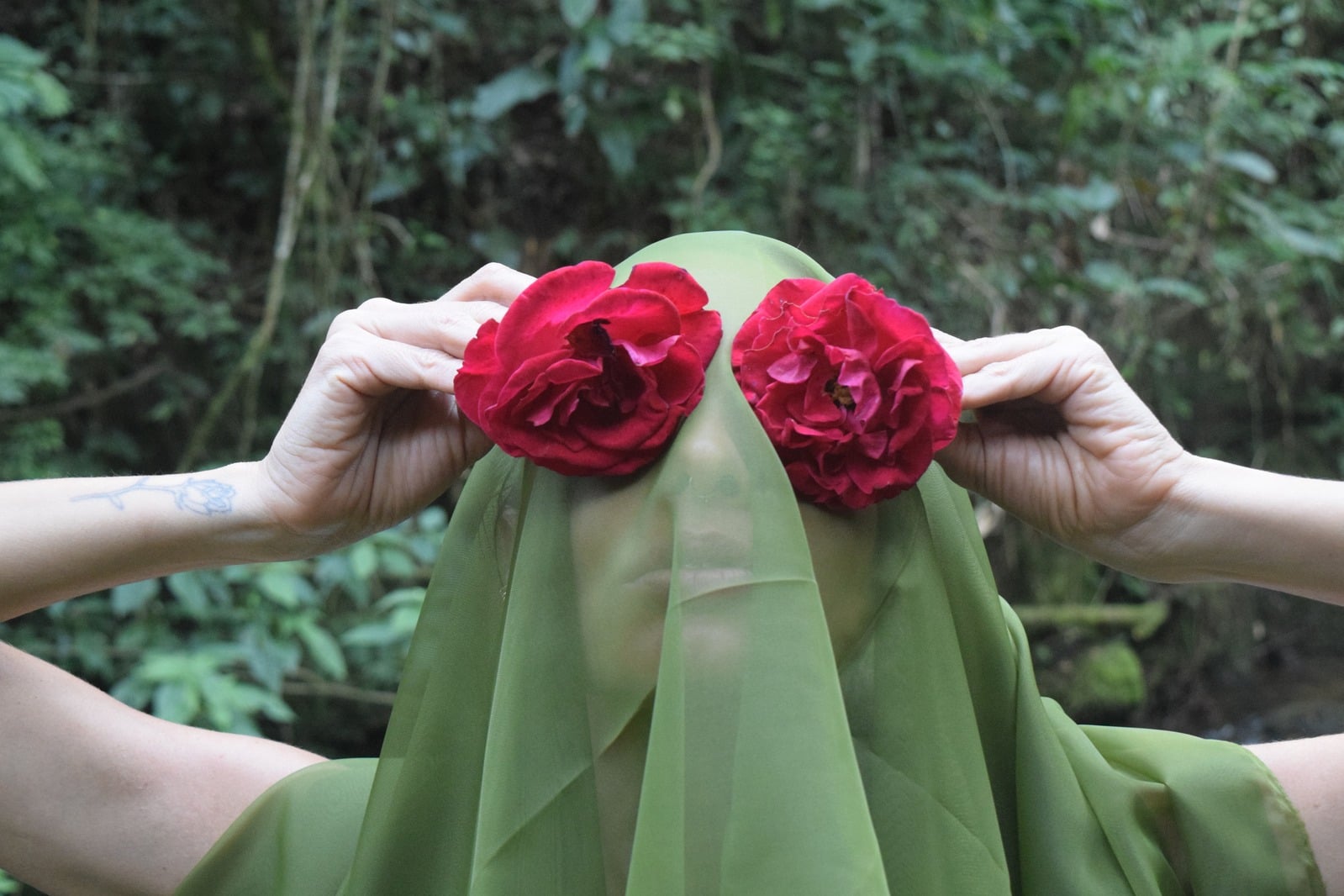 Duas mãos seguram tecidos verdes com rosas vermelhas diante da mata