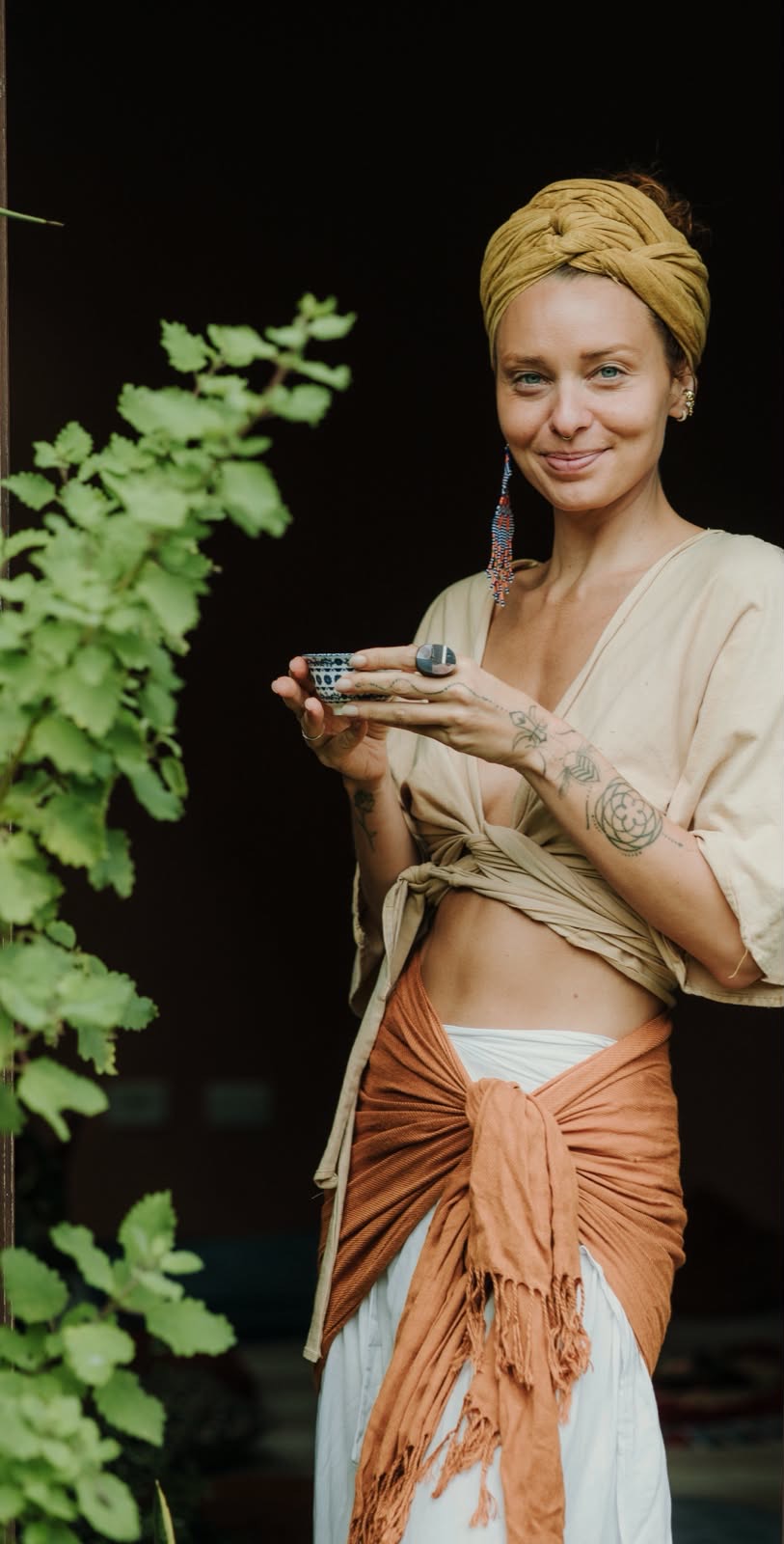 Victoria Pastorello com turbante dourado, segurando uma xícara junto às plantas